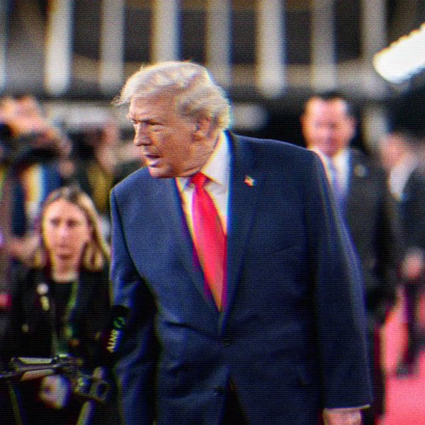 Donald Trump on a red carpet with lights and press cameras around him. He leans in talking to the press.