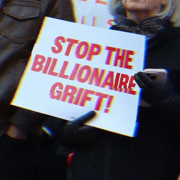 Two protestors, one holds a sign reading ‘stop the billionaire grift,’ the other reads ‘protect consumers not billionaires.’