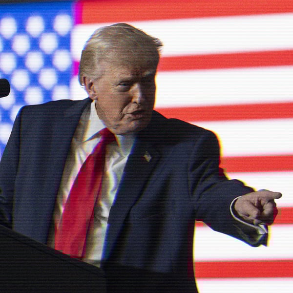 Donald Trump speaking at a podium with an American flag lit behind him. He points out at the audience.
