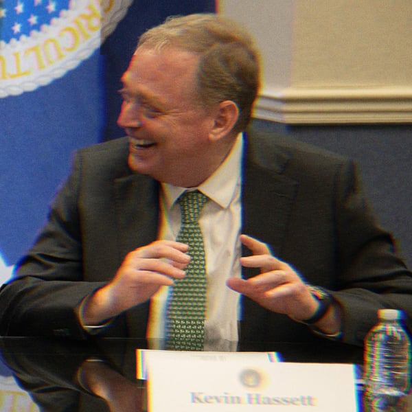 Kevin Hassett in a meeting, laughing. His name placard is visible on the table in front of him.