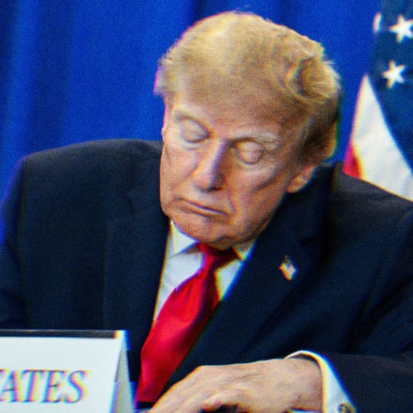 Trump at a ceremony, sitting at a table behind a placard reading, 'United States.' He is either looking down or sleeping.