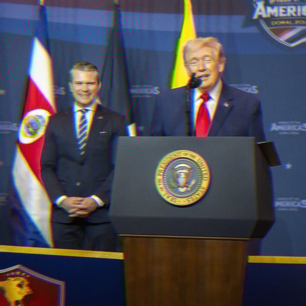 Trump is speaking at a podium. Pete Hegseth stands behind him, smiling.