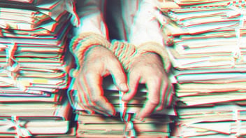 Glitchy photo of three stacks of paperwork. A man's wrists are bound by rope handcuffs and rest on the middle stack of papers. 