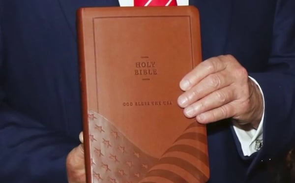 Trump holding a King James bible with the American flag and “God Bless the USA” inscribed on the cover.
