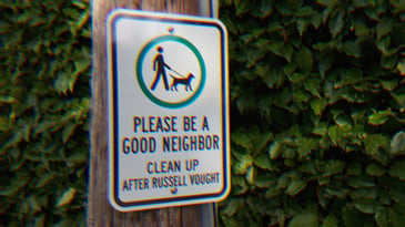 A metal sign on a pole in a park; An icon of a person walking a dog with text that reads, ‘Please be a good neighbor. Clean up after Russell Vought.’