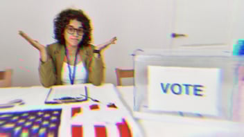 A person sitting in front of an American flag at a polling check in table shrugging, confused, with a Vote box in front of them