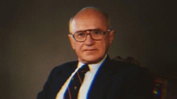 A portrait of Milton Friedman. He wears thick, round glasses, a blue suit jacket and red and black striped tie.