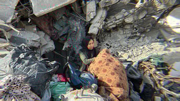 A woman in Gaza sorts through rubble; all that's left of her home. She is surrounded by crumbling concrete and drywall, along with miscellaneous clothing and fabric.