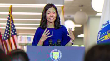 Boston Mayor Michelle Wu speaking to a crowd, standing at a podium with American flags behind her.