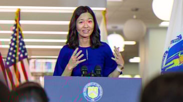 Boston Mayor Michelle Wu speaking to a crowd, standing at a podium with American flags behind her.