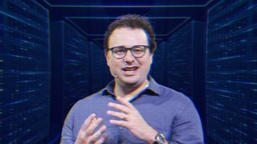Dario Amodei mid-speech, he's wearing a wireless mic and is talking with his hands. His image is overlayed over a rendering of futuristic server room.