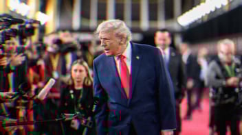 Donald Trump on a red carpet with lights and press cameras around him. He leans in talking to the press.