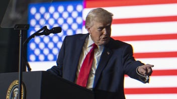 Donald Trump speaking at a podium with an American flag lit behind him. He points out at the audience.