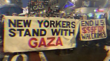 Protestors holding banners. One reads, ‘New Yorkers Stand with Gaza.’ Another, ‘End U.S. Money for War Crimes.’ Posters behind read, ‘Save Gaza,’ and ‘Defund the Genocide.’