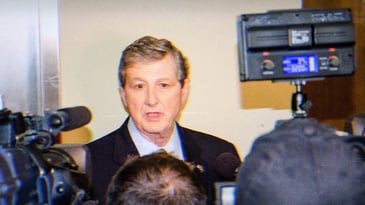 Senator John Kennedy speaking at a press conference. There are cameras and microphones in his face and he has a goofy expression on his face.