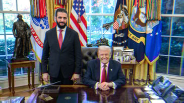 Syrian President Ahmed al-Sharaa with Donald Trump in the Oval Office. al-Sharaa stands next to Trump who is sitting.