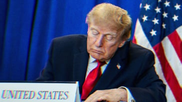Trump at a ceremony, sitting at a table behind a placard reading, ‘United States.’ He is either looking down or sleeping.