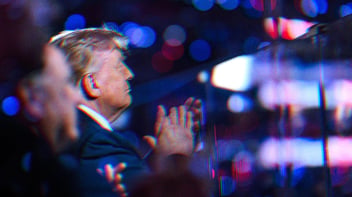 Trump at an event, red and blue lights dot the background. He is clapping and standing behind a glass barrier.