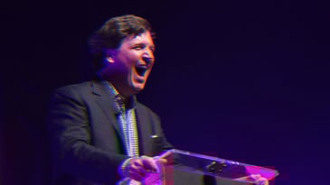 Tucker Carlson standing at a podium mid laugh, smiling.