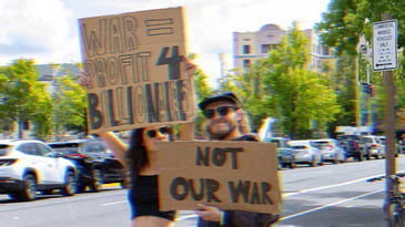 Two people holding protest signs. One reads, ‘War= Profit 4 Billionaires,’ the other ‘Not Our War.’