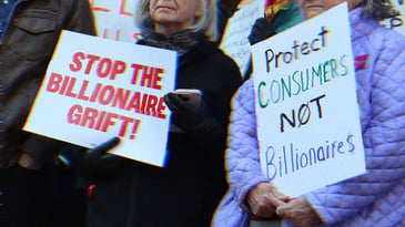 Two protestors, one holds a sign reading ‘stop the billionaire grift,’ the other reads ‘protect consumers not billionaires.’