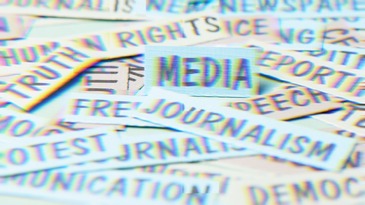 A pile of paper strips bearing words associated with press freedom and civic life, including “Media,” “Journalism,” “Truth,” “Rights,” “Free,” “Speech,” “Protest,” “Democracy,” “Newspaper,” “Reporting,” and “Communication,” with the word “Media” centered on a small card in the foreground.