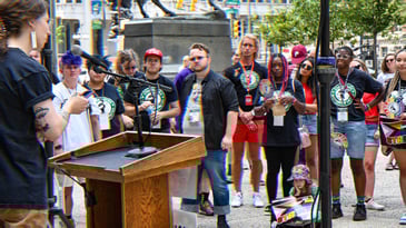 Starbucks workers and organizers gathered at City Hall in Philadelphia. Someone stands at a podium speaking to the group that is gathered. Many are wearing shirts with the Starbucks Workers United logo on them.