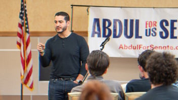 U.S. Senate candidate for Michigan Abdul El-Sayed talks to a crowd. An ‘Abdul for U.S. Senate’ banner appears behind him, as well as an American flag.