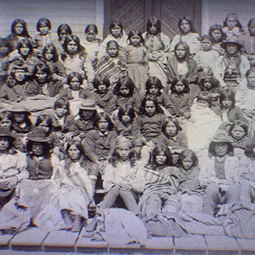 Ciricahua Apaches at the Carlisle Indian School. Children and adults sit on the porch of the school.
