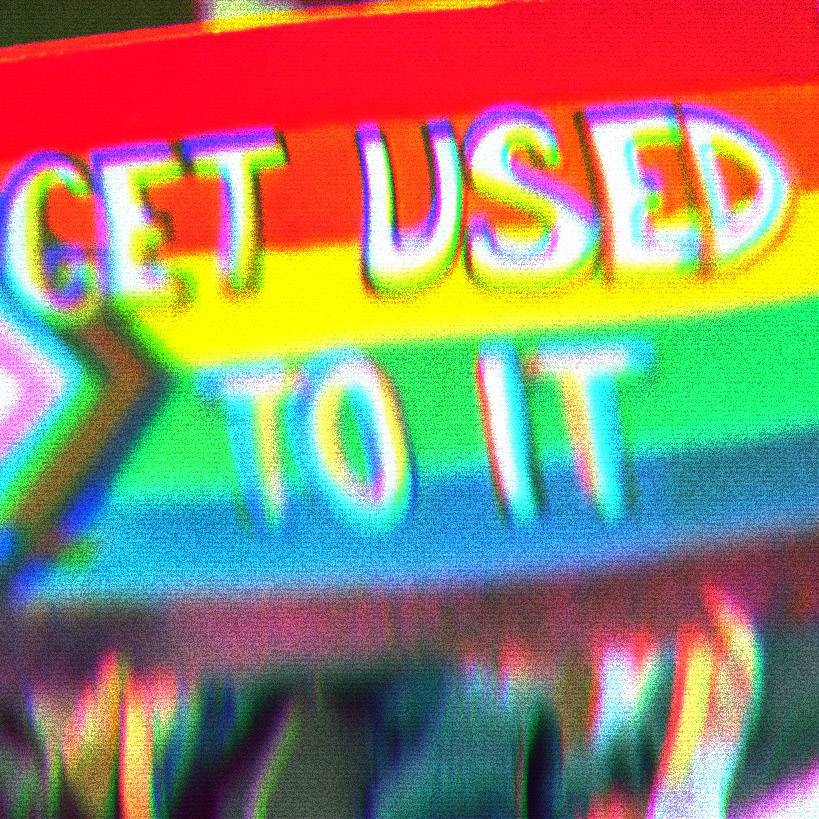 Two people holding a sign that says 'Get Used To It.' The sign background is the modern pride flag