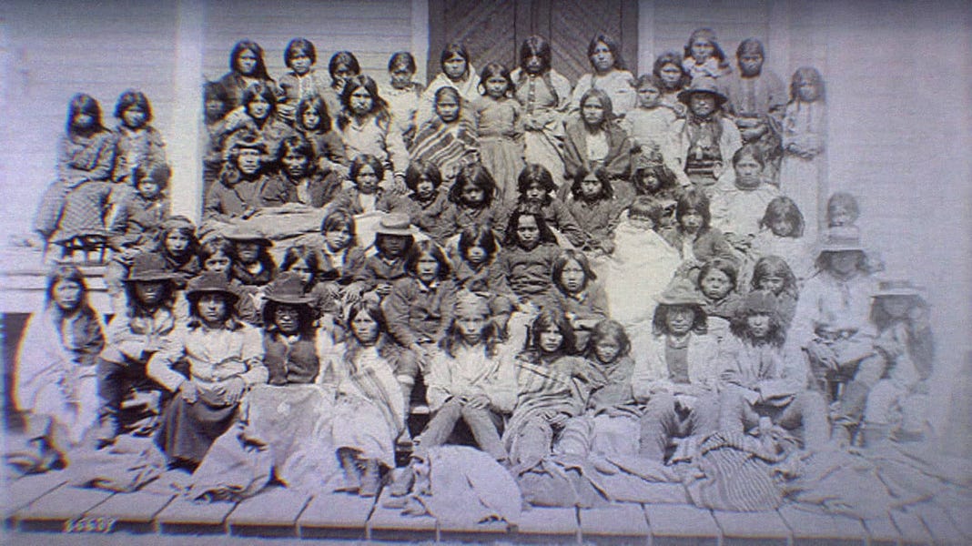 Ciricahua Apaches at the Carlisle Indian School. Children and adults sit on the porch of the school. 