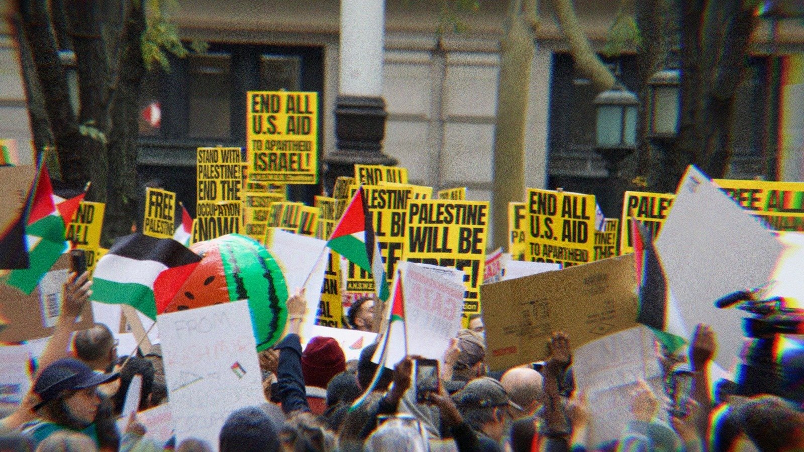 A group of Pro-Palestine protestors in NYC. Palestinian flags and signs are held up with messages like 'End all U.S. aid to apartheid Israel,' and 'Palestine will be free.'