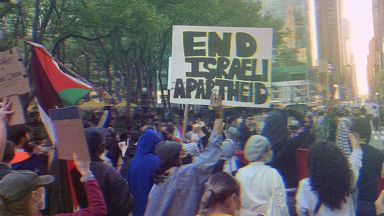 A large group of protesters gathering in NYC. The Palestinian flag is visible, as well as a sign that reads ‘End Israeli Apartheid.’