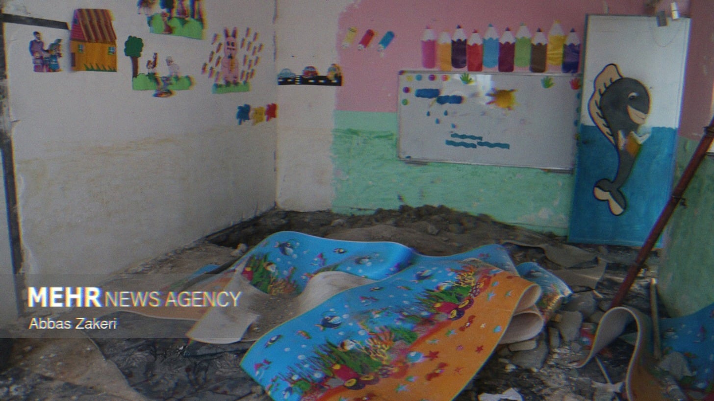 A room in the Shajareh Tayyebeh school. Rubble lines the floor, along with damaged banners depecting marine life. Decorations like farm animals, colored pencils, and a whiteboard line the walls. A once vibrant classroom now condemned