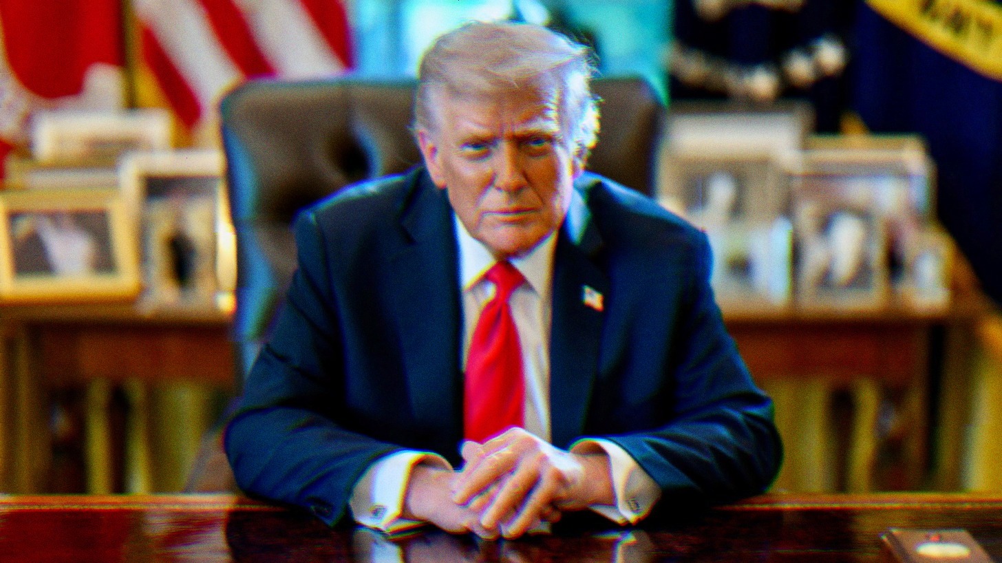 Donald Trump sitting at his desk. He is wearing a blue suit, a red tie, and has his hands on the desk, one on top of the other. He is attempting to make a tough-looking, intimidating facial expression.