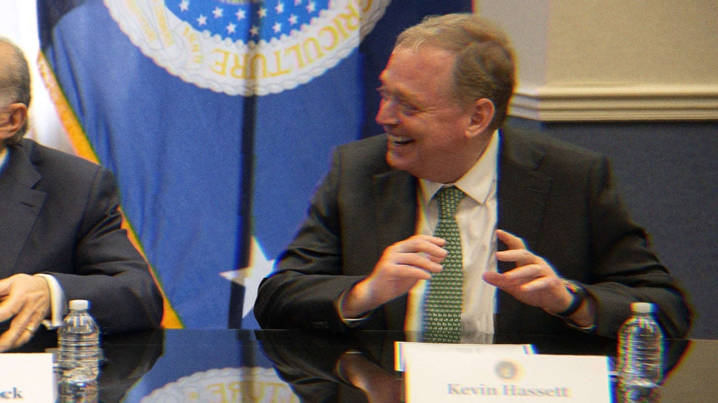 Kevin Hassett in a meeting, laughing. His name placard is visible on the table in front of him.
