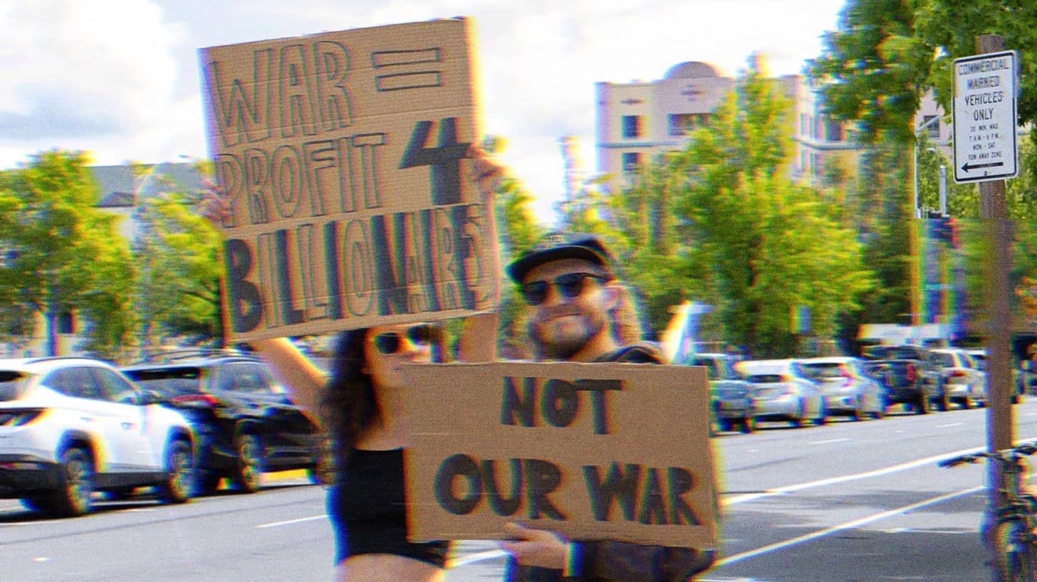 Two people holding protest signs. One reads, ‘War= Profit 4 Billionaires,’ the other ‘Not Our War.’