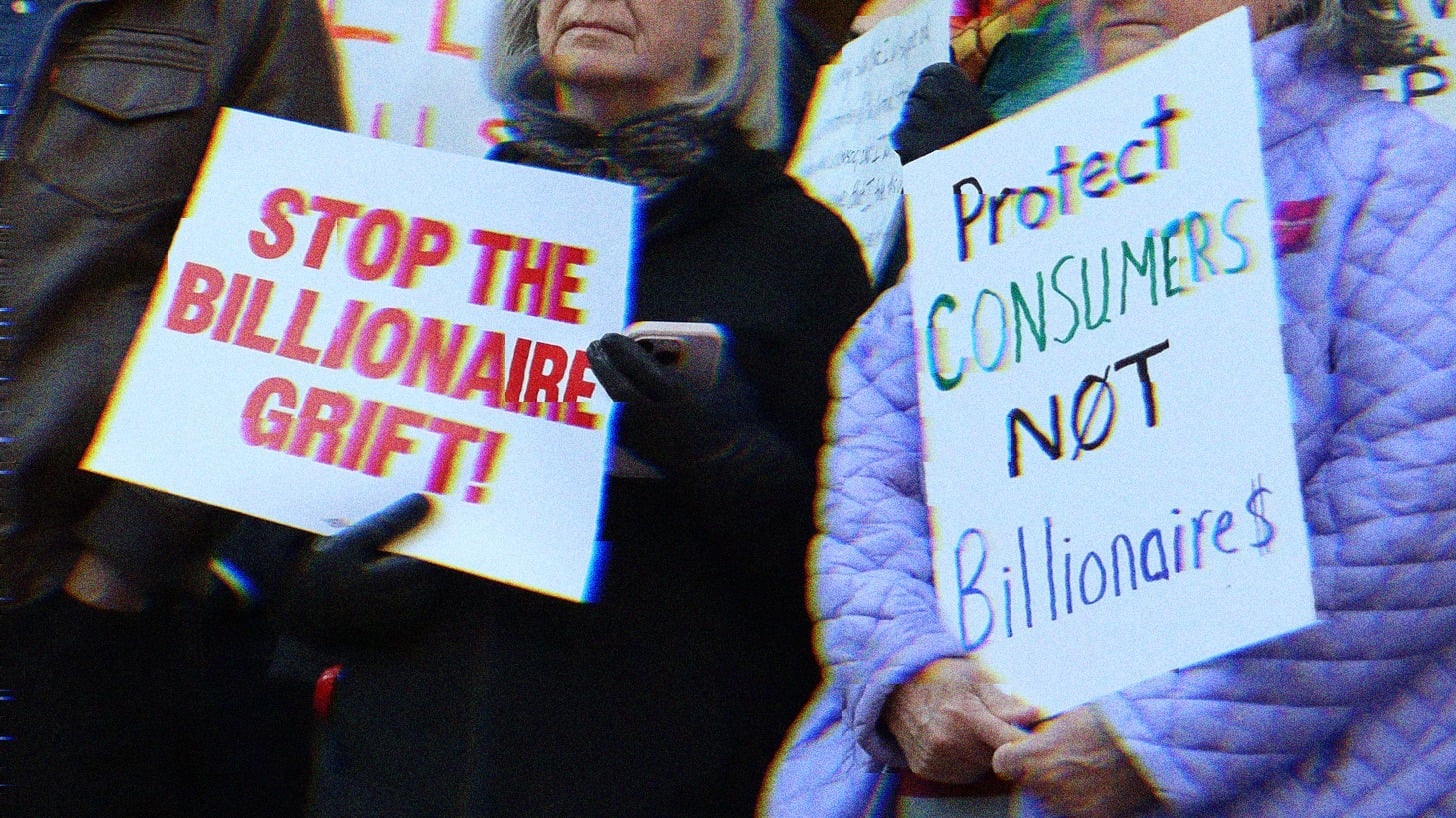 Two protestors, one holds a sign reading ‘stop the billionaire grift,’ the other reads ‘protect consumers not billionaires.’