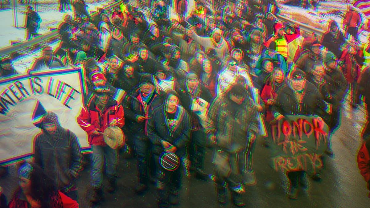 Water Protectors protesting the Dakota Access Pipeline in 2017. A man is holding a sign that says, Honor The Treatys, another says, Water Is Life.